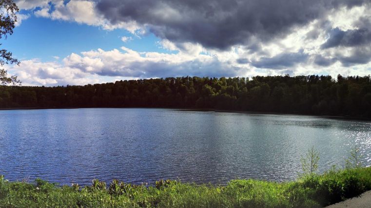 Ein ruhiger See umgeben von Bäumen und einer grünen Wiese. Der Himmel ist teilweise bewölkt mit Strahlen von Sonnenlicht.