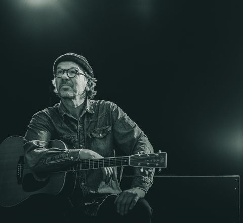 A thoughtful musician sits with a guitar. The scene is in black and white and conveys a creative atmosphere.