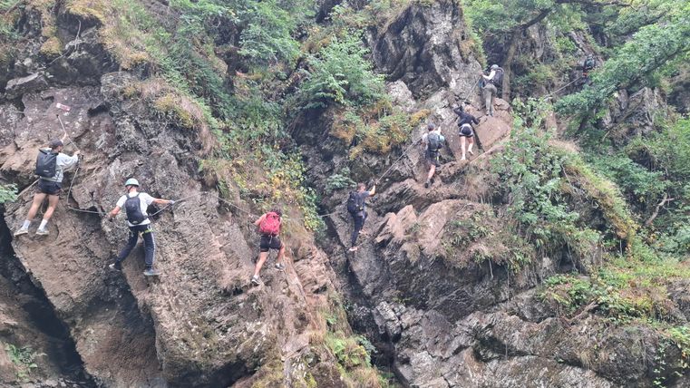 Eine Gruppe von Menschen klettert an steilen Felsen entlang. Die Umgebung ist grün und bewaldet, mit einer Gewässerstelle in der Nähe.