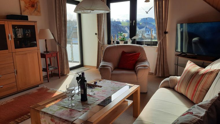 Ein gemütliches Wohnzimmer mit einem hellen Sofa und einem Sessel. Große Fenster bieten einen Blick auf die Landschaft.