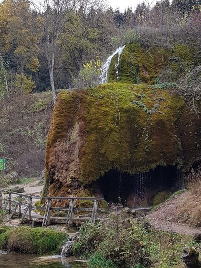Ein kleiner Wasserfall fließt über moosbedeckte Felsen. Im Vordergrund ist ein ruhiger Fluss und eine Holzbrücke zu sehen.
