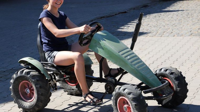 Ein Mädchen sitzt auf einem grünen Pedal-Gokart. Es ist sonnig und die Umgebung ist freundlich.