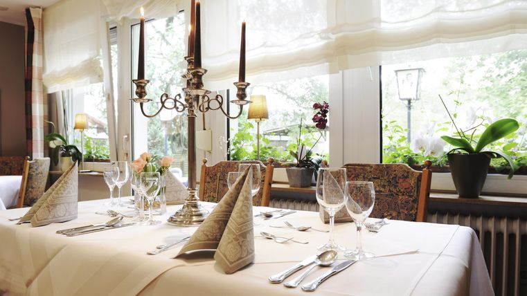 Ein elegant gedeckter Tisch mit Kerzen und Gläsern. Die Fenster lassen viel Licht herein und umgeben den Tisch mit Pflanzen.