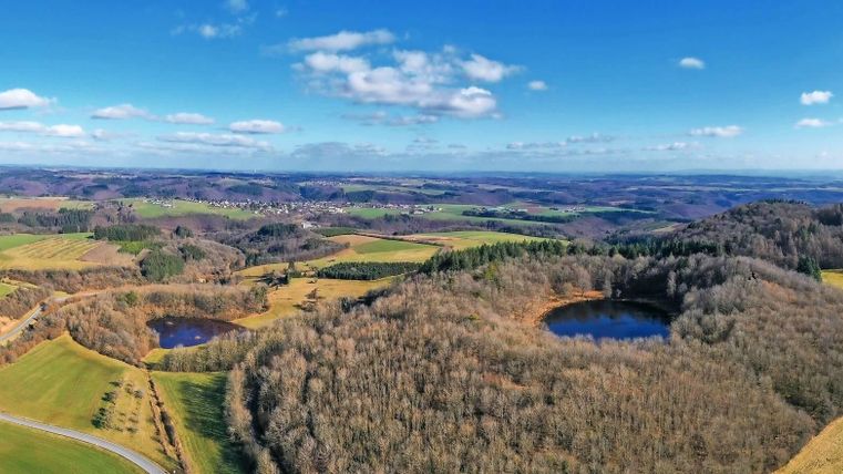 Eine malerische Landschaft mit sanften Hügeln und bewaldeten Bereichen. Im Vordergrund sind zwei runde Gewässer sichtbar, umgeben von Wiesen und Bäumen.