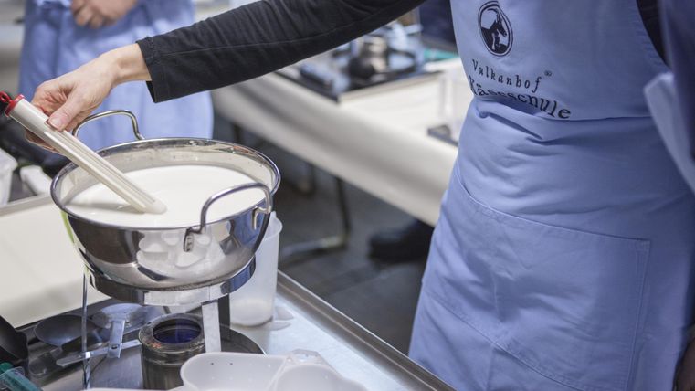Eine Person in einer blauen Schürze bereitet Milch in einer Schüssel vor. Im Hintergrund sind Kochutensilien und weitere Personen zu sehen.