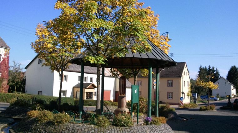 Ein hübscher Pavillon umgeben von bunten Herbstbäumen in einer ruhigen Straße. Im Hintergrund sind alte Häuser und ein klarer blauer Himmel zu sehen.