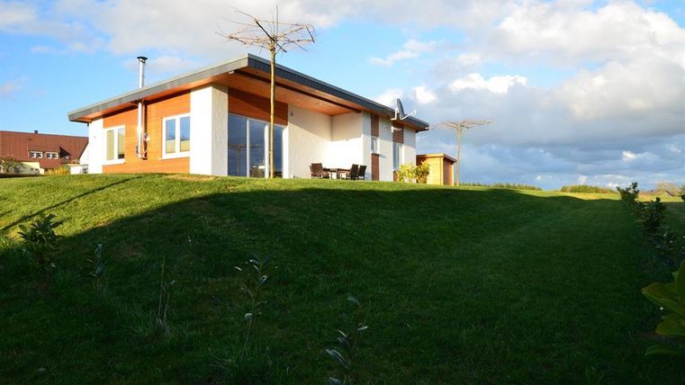 Ein modernes Haus auf einer sanften Erhöhung mit grünem Rasen. Der Himmel ist teilweise bewölkt und es sind einige Möbel auf der Terrasse sichtbar.
