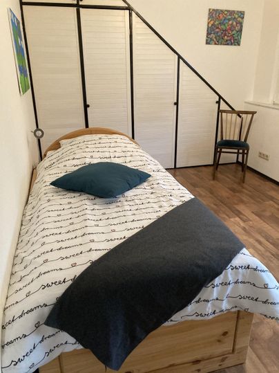 Ein schlichtes Schlafzimmer mit einem Bett, einer Decke und einem Kissen. Im Hintergrund befindet sich ein Stuhl und eine leichte Treppe.