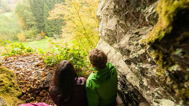 Ein Blick auf eine malerische Landschaft mit buntem Herbstlaub. Zwei Personen sitzen in einer Höhle und genießen die Aussicht.