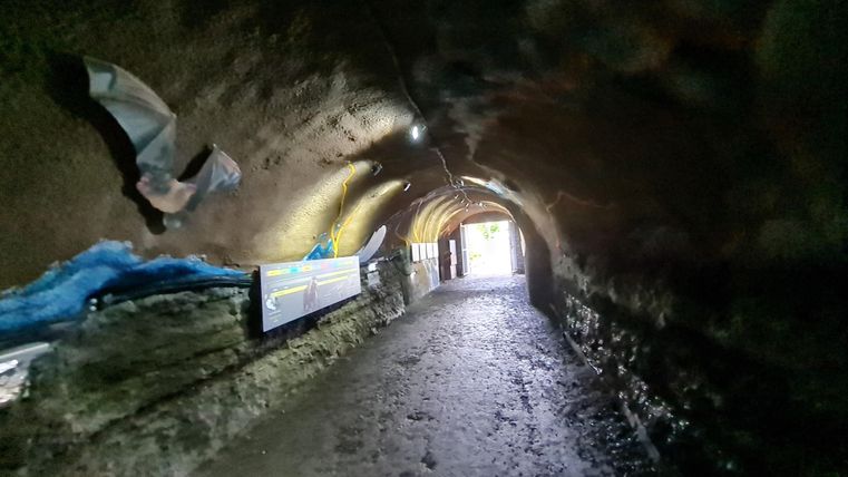 Ein langes, gewölbtes Tunnel mit feuchten Wänden und einem klaren Ausgang am Ende. An den Seiten sind einige Tafeln angebracht.