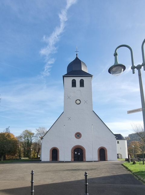 Ein ruhiger Bach fließt durch eine grüne Wiese mit Bäumen im Hintergrund. Der Himmel ist bewölkt und die Landschaft wirkt friedlich.