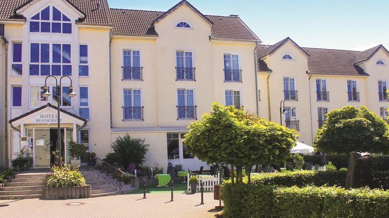 Ein modernes Hotelgebäude mit großen Fenstern und einem einladenden Eingangsbereich. Umgeben von gepflegten Gärten und Sitzgelegenheiten im Freien.