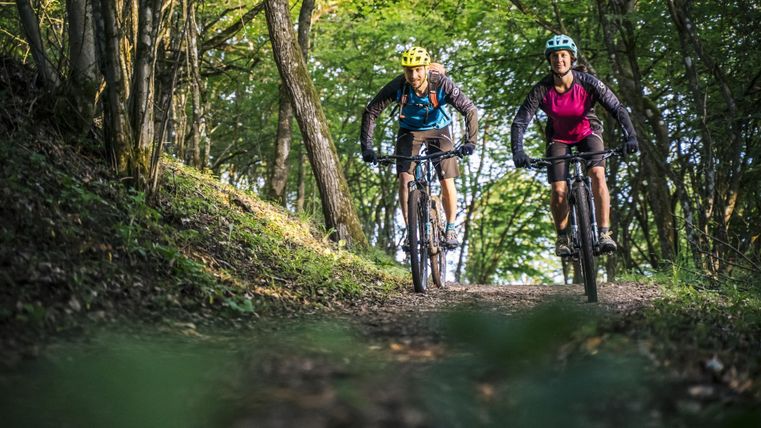 Zwei Radfahrer auf einem Waldweg, umgeben von grünen Bäumen. Sie tragen Helme und sehen entspannt aus.