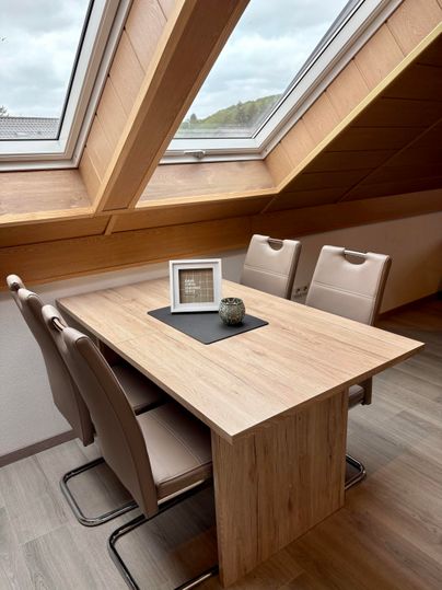 A bright attic room with a wooden dining table and four chairs. Large windows offer a view of the landscape.