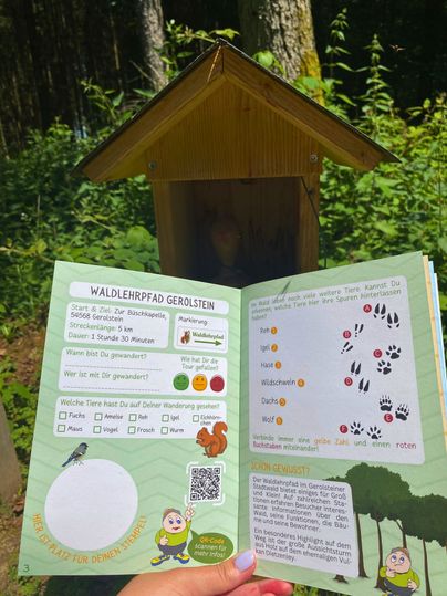 Die zwei Seiten zum Waldlehrpfad im Wander(S)pass für Kids werden offen gezeigt. Dahinter befidnet sich die zugehörige Stempelstation, worin sich der Stempel für den Pass befindet.