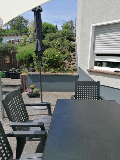 Ein gemütlicher Außenbereich mit einem Tisch und Stühlen. Im Hintergrund sind Pflanzen und ein Sonnenschirm zu sehen.