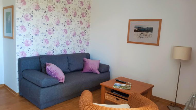 Ein gemütliches Wohnzimmer mit einem grauen Sofa und einem Korbstuhl. Die Wände sind mit blühendem Hintergrundmuster dekoriert.