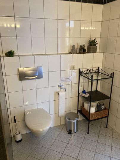 Ein modernes Badezimmer mit weißen Fliesen. Es enthält eine Toilette, eine Papierhalterung und ein Regal mit Handtüchern und Pflanzen.