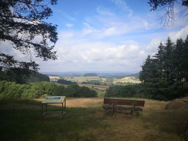 Ein schöner Ausblick über das Tal mit sanften Hügeln und bewaldeten Flächen. Im Vordergrund stehen eine Bank und ein Informationstisch.