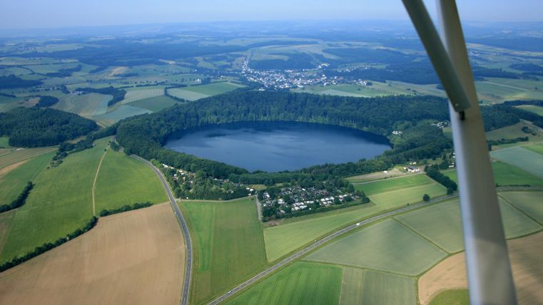 Eine Landschaft mit einem runden See, umgeben von grünen Feldern und Wäldern. Teilweise bewaldete Hügel und ein kleines Dorf sind ebenfalls sichtbar.