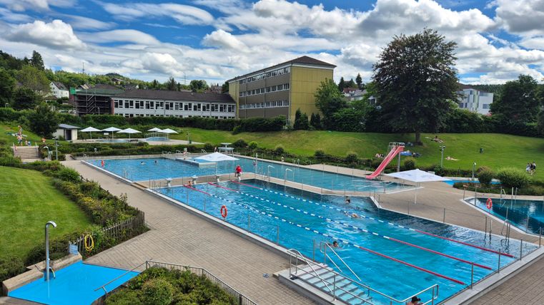 Ein schöner Freibadbereich mit mehreren Becken und Wasserrutschen. Umgeben von grünen Wiesen und Bäumen unter einem blauen Himmel.