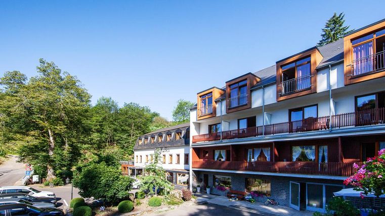 Ein modernes Hotelgebäude umgeben von Bäumen und Grünflächen. Auf dem Gelände sind parkende Autos sowie Balkone mit Blick ins Freie zu sehen.