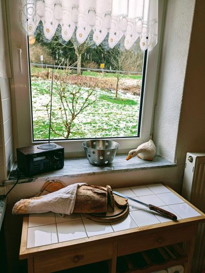 Eine gemütliche Küchenecke mit einem Fenster, durch das man einen winterlichen Garten sehen kann. Auf dem Tisch liegen ein Brotmesser, ein Laib Brot und eine Tischdecke.