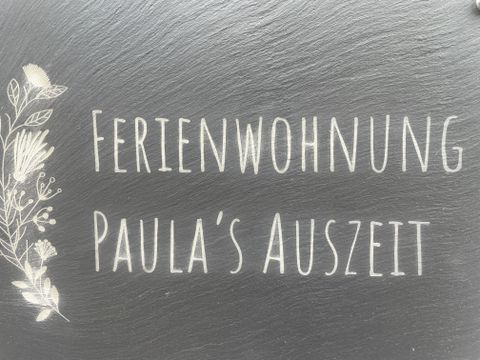 Ein Schild mit der Aufschrift "Ferienwohnung Paula's Auszeit". Es zeigt eine einladende Atmosphäre für Urlaubsgäste.