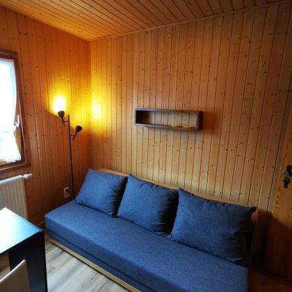 Ein gemütlicher Raum mit holzverkleideten Wänden. Ein blauer Sofa mit Kissen und eine Stehlampe schaffen eine einladende Atmosphäre.