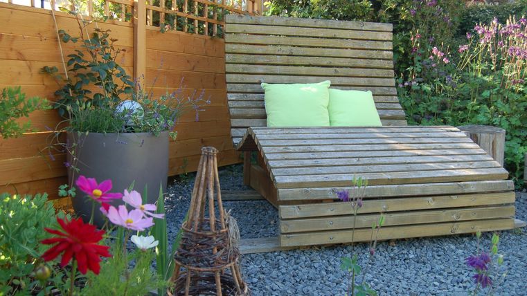 Eine gemütliche Liege aus Holz steht in einem blühenden Garten. Bunte Blumen und eine schöne Gartenstruktur umgeben den Relaxbereich.