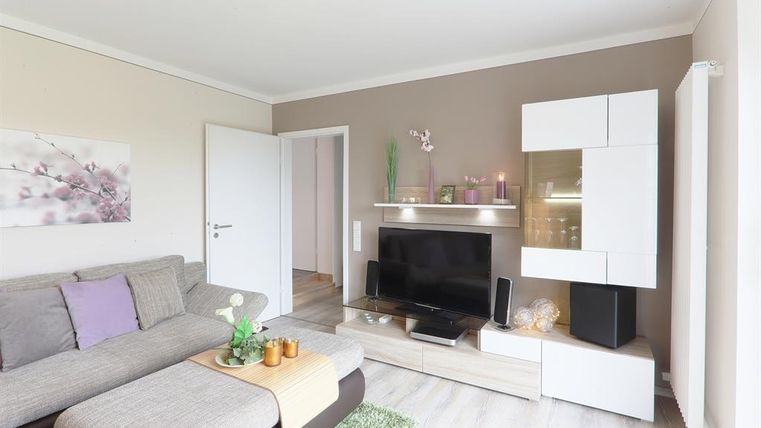 Ein modernes Wohnzimmer mit einem grauen Sofa und einem Fernseher auf einem hellen TV-Möbel. Die Wände sind in neutralen Farben gehalten, und es gibt dekorative Elemente wie Pflanzen und einen Teppich.