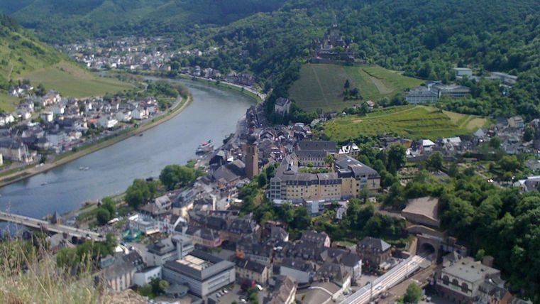 Eine malerische Stadt am Fluss mit grünen Hügeln im Hintergrund. Die Architektur besteht aus traditionellen Gebäuden, die sich harmonisch in die Landschaft einfügen.