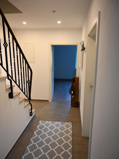 Ein heller Flur mit einer Treppe und einem dekorativen Teppich. Im Hintergrund ist eine offene Tür, die zu einem weiteren Raum führt.
