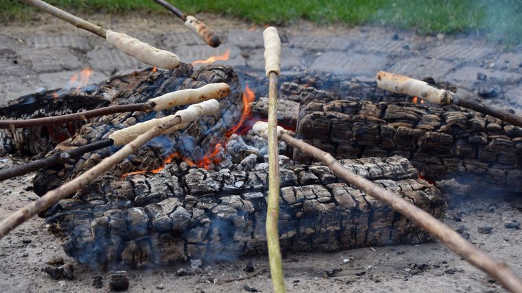 Ein Lagerfeuer mit verbrannten Holzscheiten liegt im Mittelpunkt. Stöcke mit Teig sind über die Flammen gehalten.