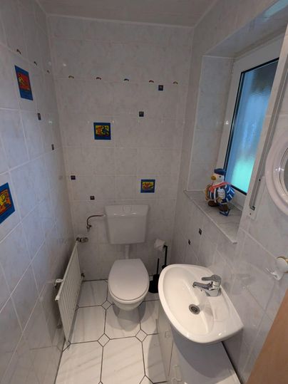 Ein kleines, modernes Badezimmer mit weißen Fliesen. Es enthält ein Waschbecken, eine Toilette und ein Fenster.