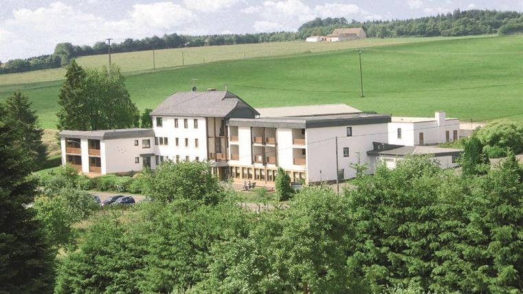 Ein modernes Gebäude umgeben von Bäumen und Wiesen. Der Himmel ist klar und die Landschaft wirkt friedlich.