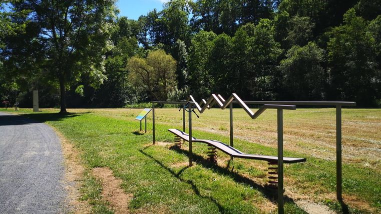 Ein Fitnessbereich im Freien mit Turngeräten auf einer grünen Wiese. Im Hintergrund sind Bäume und ein blauer Himmel zu sehen.