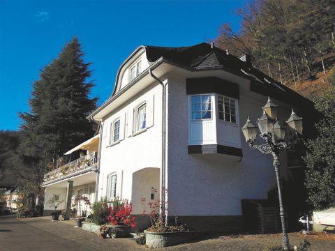 Ein charmantes Haus mit zwei Etagen und einem Balkon, umgeben von Bäumen und Blumen. Die Fassade ist weiß, und eine alte Laterne steht vor dem Gebäude.