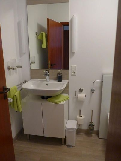 Ein modernes Badezimmer mit einem Waschbecken, einem Spiegel und einem Holzschrank. Die Wände sind hell, und es gibt grüne Handtücher sowie Toilettenpapierhalter.