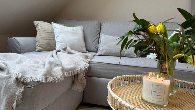 Ein gemütliches Wohnzimmer mit einem grauen Sofa und Kissen. Auf dem Tisch steht eine Vase mit frischen Blumen und eine Kerze.