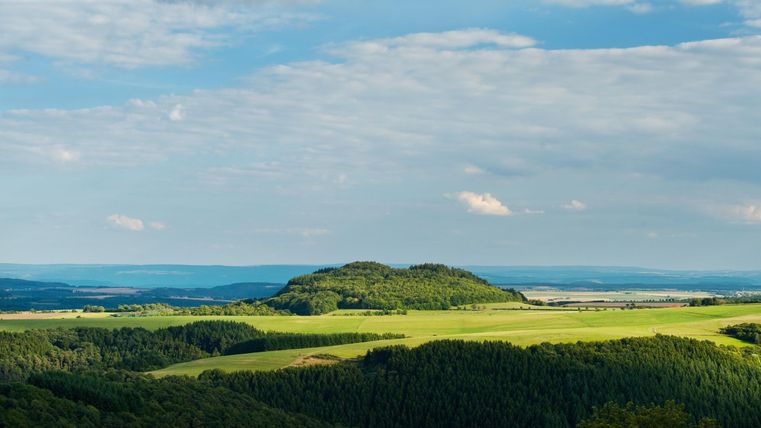 Eine weite Landschaft mit sanften Hügeln und viel Grün. Der Himmel ist bewölkt, aber es gibt auch klare Stellen.
