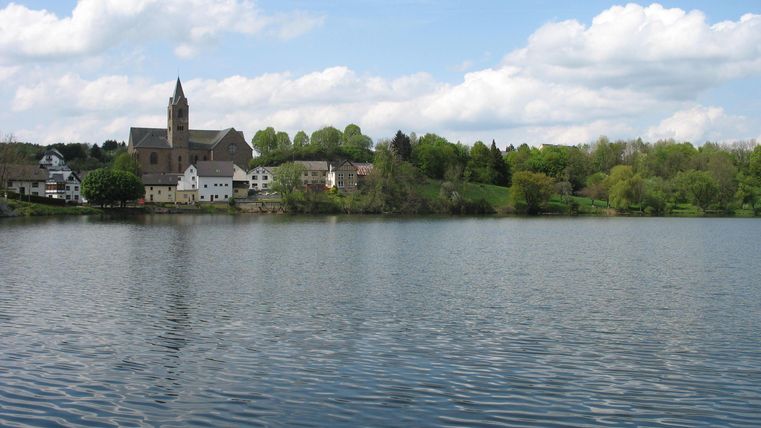 Ein ruhiger See mit klarer Wasseroberfläche. Im Hintergrund sind eine Kirche und einige Häuser umgeben von Bäumen und blauem Himmel.