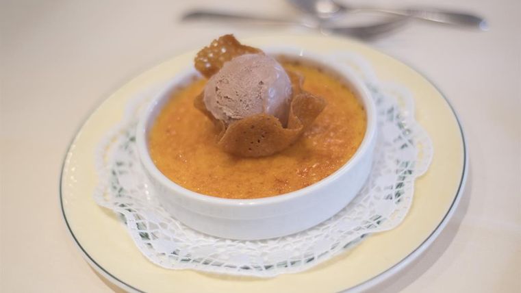 Eine köstliche Crème brûlée mit einer Schokoladeneiskugel und einem knusprigen Waffelblatt. Das Dessert ist in einer weißen Schale auf einem grazilen Teller angerichtet.