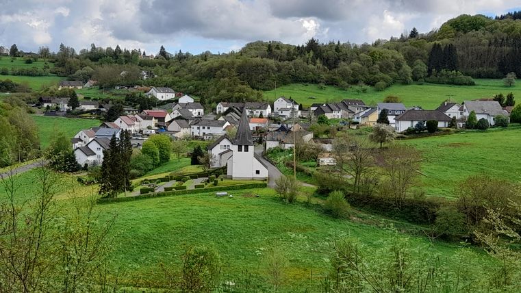 Ein malerisches Dorf in einer grünen Landschaft. Die Häuser sind umgeben von Wiesen und Bäumen, mit einer kleinen Kirche im Vordergrund.