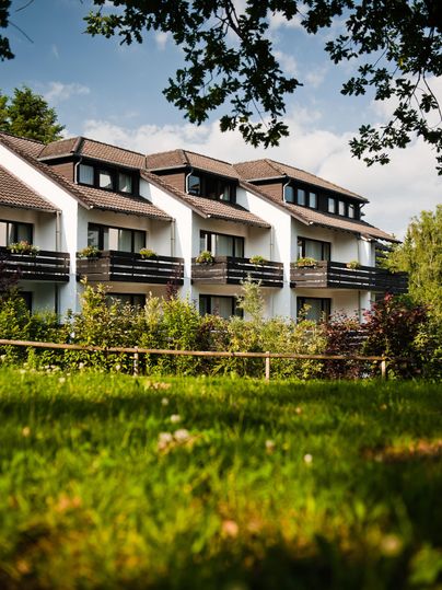 Ein modernes Gebäude mit Balkonen, umgeben von grünem Gras und Bäumen. Der Himmel ist teilweise bewölkt, was eine einladende Atmosphäre schafft.