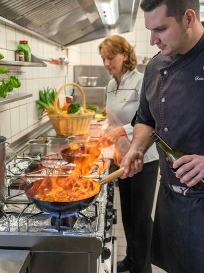In einer modernen Küche kocht ein Koch mit einem brennenden Pfanneninhalt. Eine Frau schaut lächelnd zu ihm hinüber, während frisches Gemüse im Hintergrund sichtbar ist.