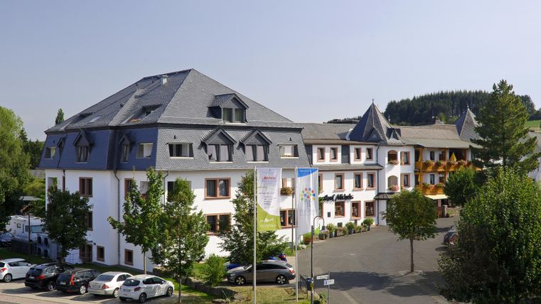 Ein modernes Hotelgebäude mit mehreren Etagen und einem gepflegten Außenbereich. Im Vordergrund sind Autos parkiert und grüne Bäume sind sichtbar.