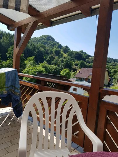 Ein Balkon mit einem weißen Stuhl und Blick auf grüne Hügel. Der Himmel ist klar und sonnig, was eine entspannte Atmosphäre schafft.