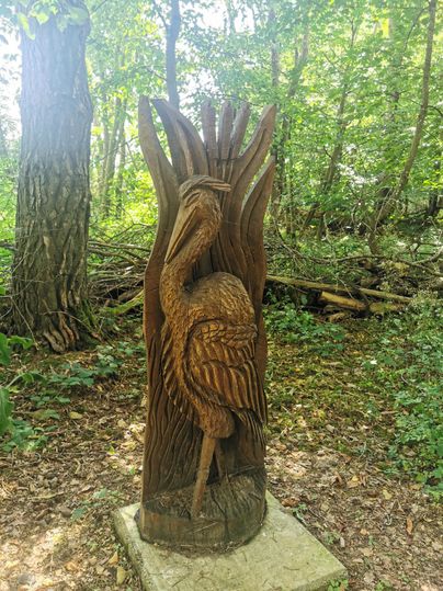 Eine schön geschnitzte Holzskulptur eines Storches steht in einem Wald. Umgeben von Bäumen strahlt das Kunstwerk eine ruhige Atmosphäre aus.