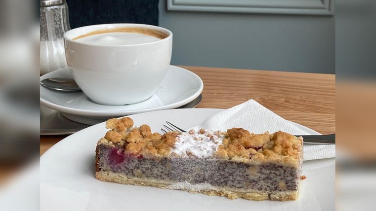 Ein Stück Kuchen mit einer crumbly Oberfläche liegt auf einem weißen Teller. Daneben steht eine Tasse Kaffee auf einem Unterteller.